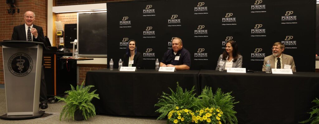A Purdue University backdrop is behind a panel of people. Two women sit on either side of a man, with plants in front of the table on the floor.