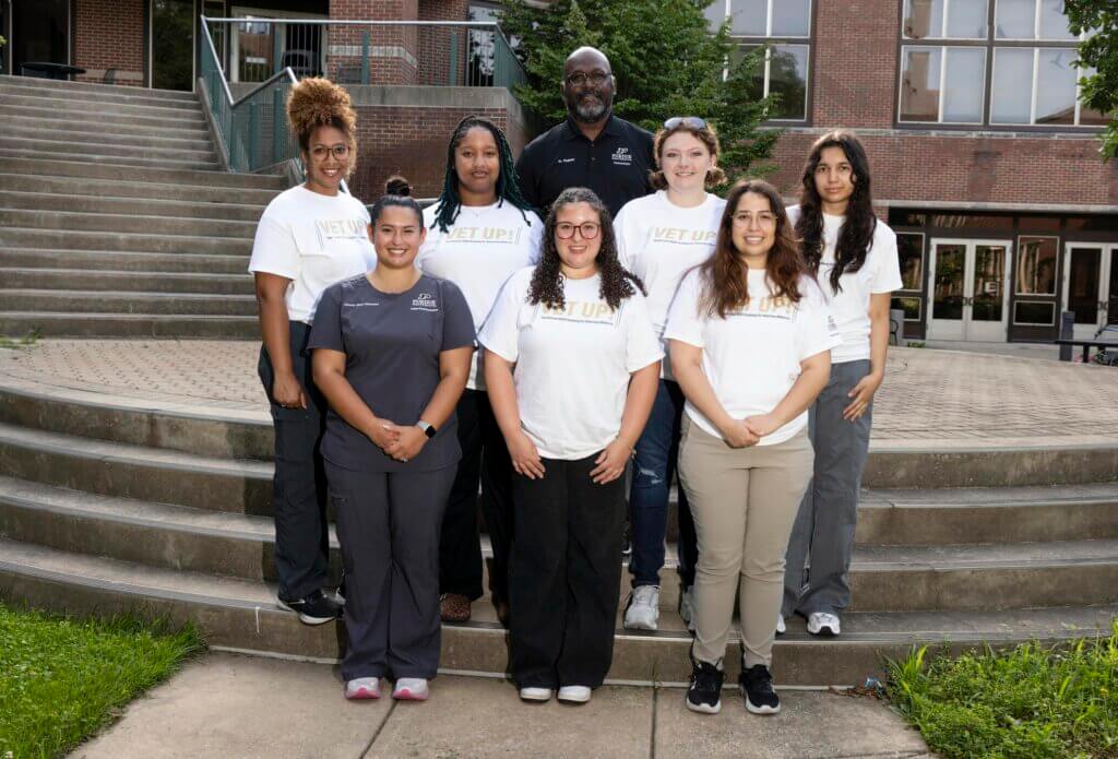 A group of people are positioned on an outdoor staircase. The majority of them are wearing white shirts that say "VET UP!" and others are wearing Purdue brand uniforms.