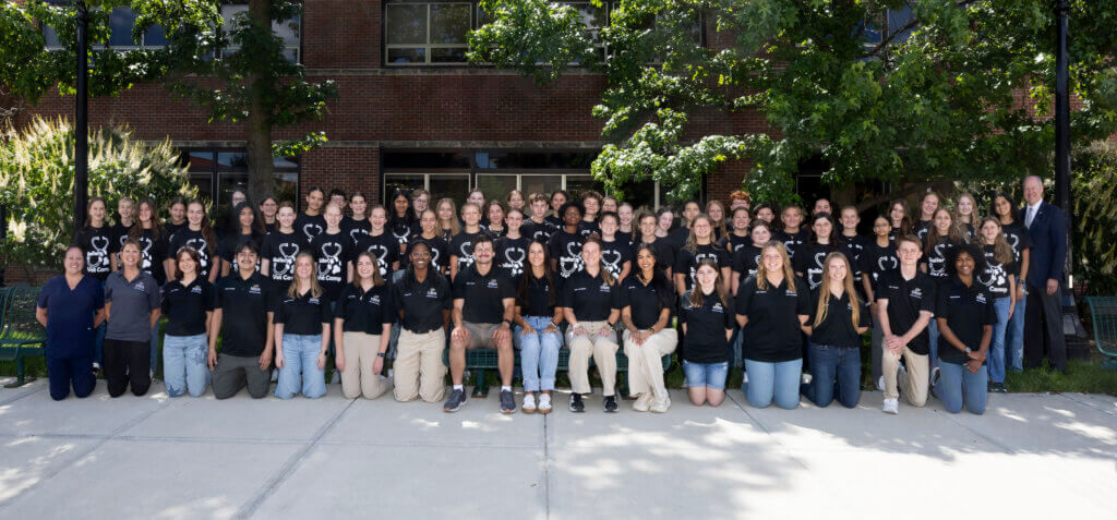 A large group of young adults and high school students are posed in an outdoor setting. All of the people are wearing black shirts.