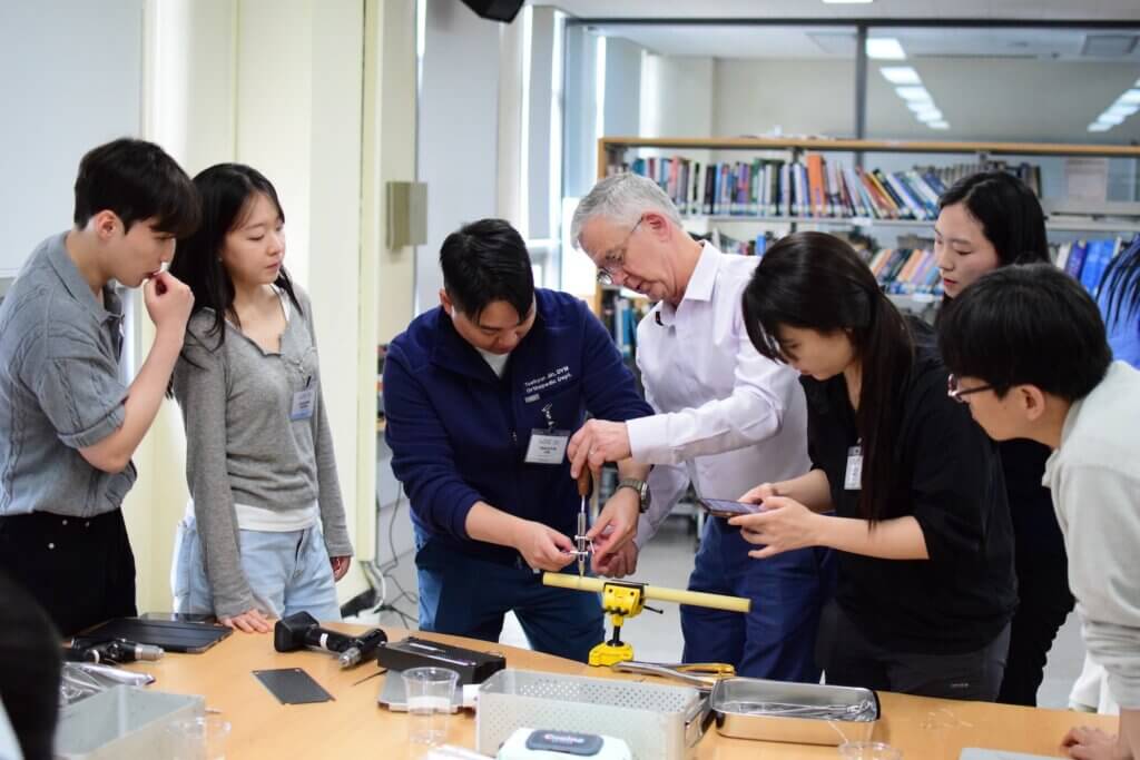 A man wearing a button up shirt is giving a hands-on demonstration to a group of six young adults in a library/classroom setting