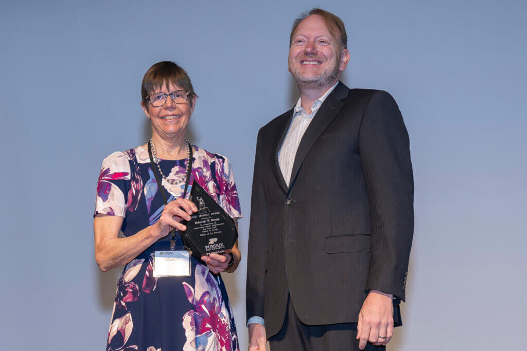 A woman wearing a floral print shirt is holding an award and is standing next to a man who is wearing a dark suit.