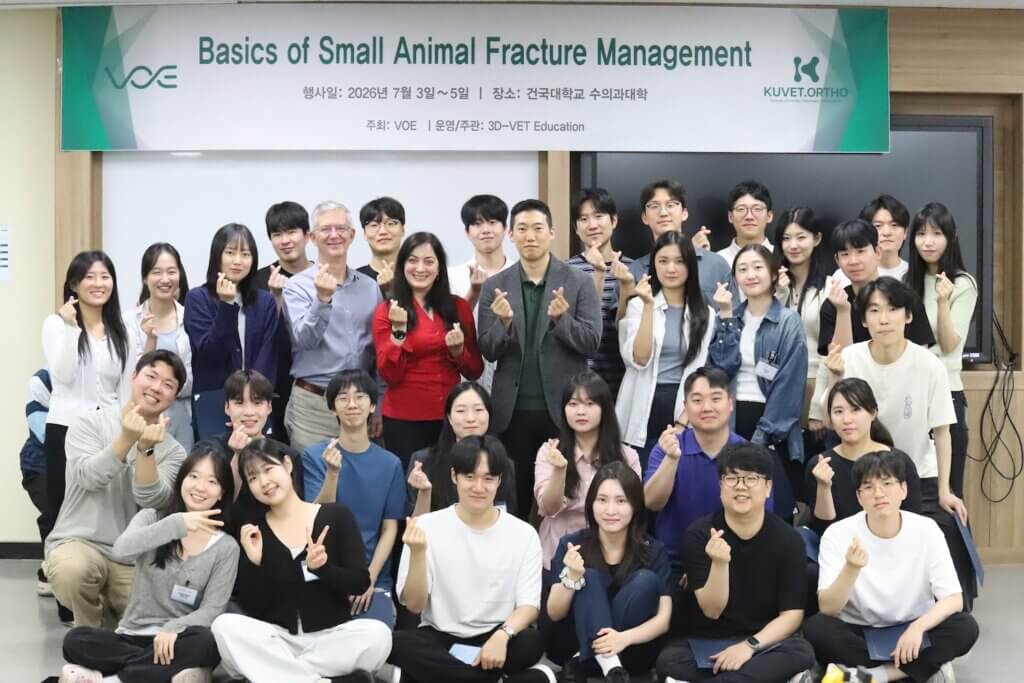 A large group of people are standing under a sign that says "Basics of Small Animal Fracture Management" and are holding up their hands to pose for a photograph.