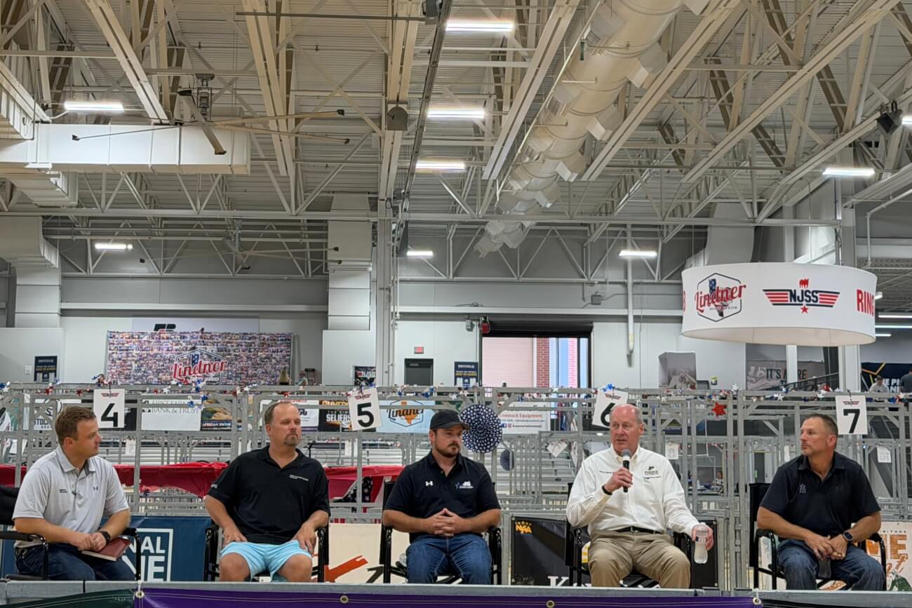 A group of men are seated in a row in what appears to be a farm animal barn. One of the men is holding a microphone will the others appear to listen attentively