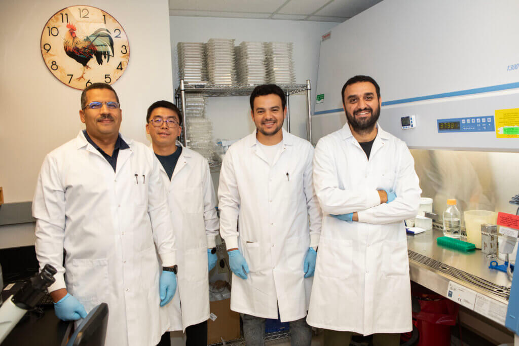 Laboratory workers are pictured in front of a fume hood and various lab equipment. The workers are wearing white coats and gloves.