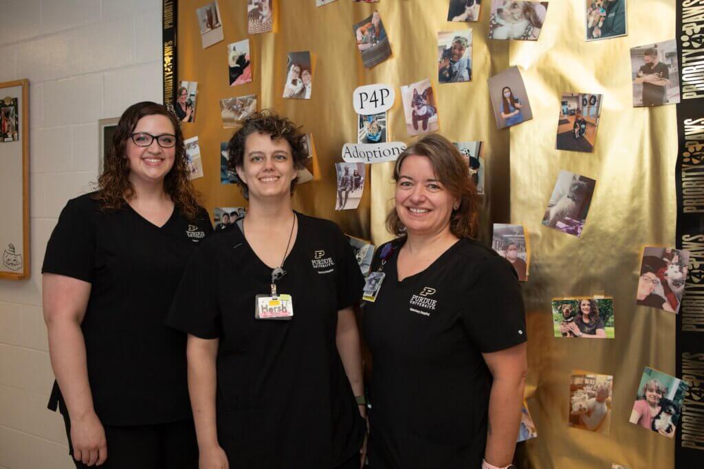 three people are standing in front of a gold backdrop with photos of animals and people on them. The people in the photograph are all wearing Purdue University Veterinary Hospital scrubs.