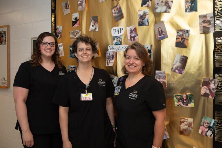 three people are standing in front of a gold backdrop with photos of animals and people on them. The people in the photograph are all wearing Purdue University Veterinary Hospital scrubs.
