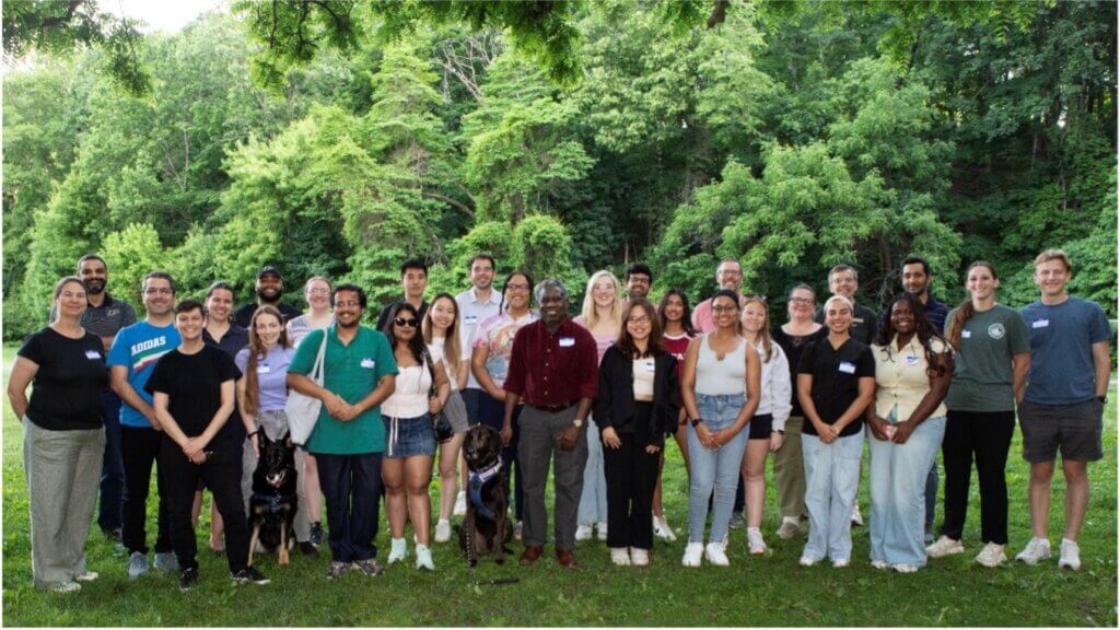 A lush, forest backdrop is behind a large group of people, all wearing casual attire.