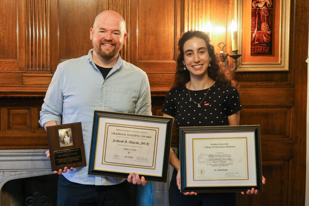 Two adults are side-by-side, holding framed award certificates. The man on the left is wearing a blue button up shirt. The woman on the right is wearing a polka dot top and a necklace