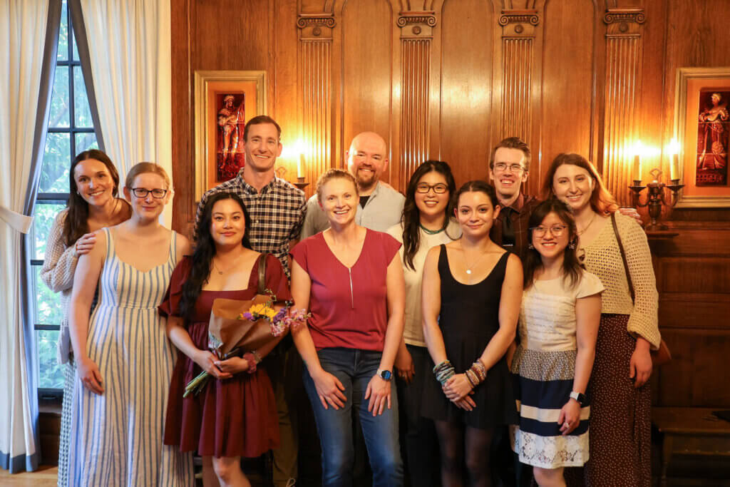 A large group of people are standing together, with one woman in the front holding a bouquet of flowers. The wall behind them is wooden and has candlesticks and framed artwork close by.