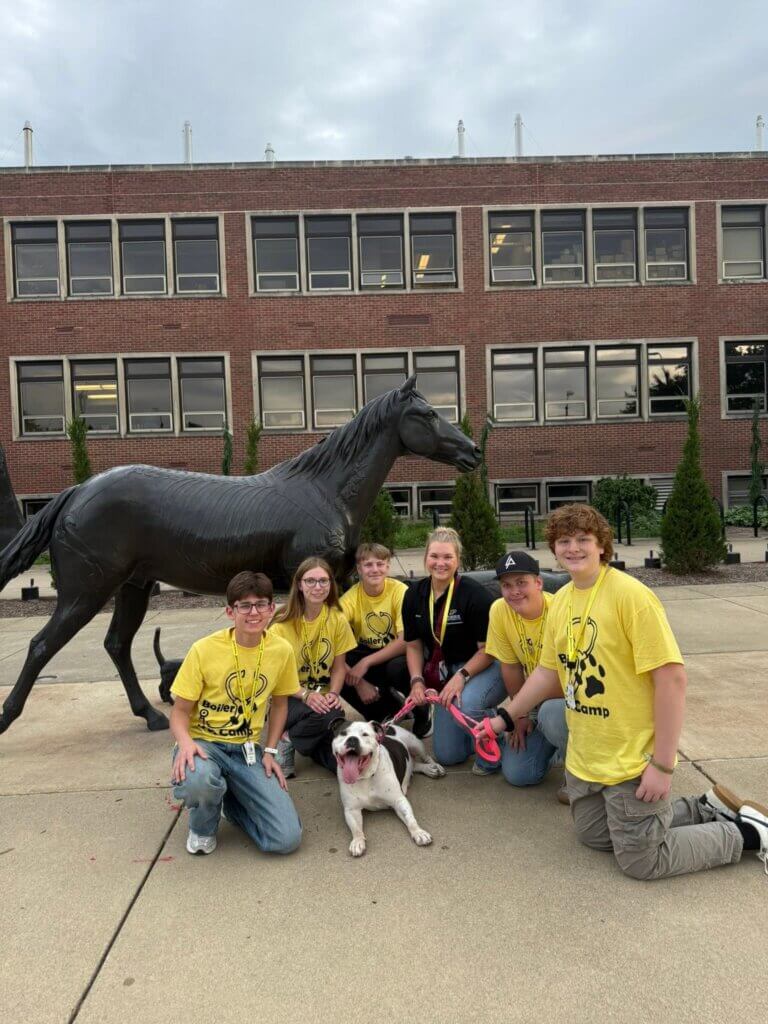 A group of teenagers are positioned with a dog. They are outside in front of a sculpted horse.