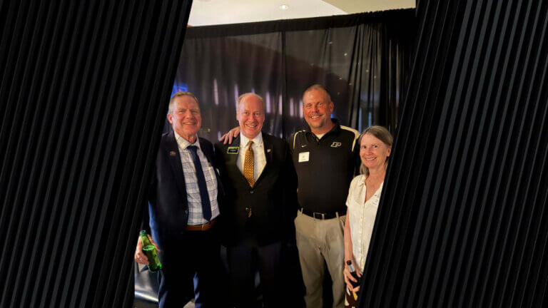 four people; three men on the left and a woman on the far right, are grouped together for a group photo at a conference. The photo is set in front of a black curtain.