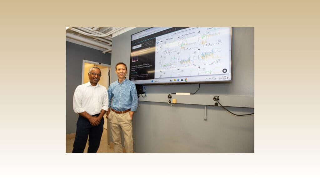 Two men are positioned on the left side of the photo next to a digital screen that shows data charts
