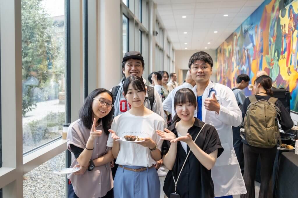 A group of five people are positioned together in a long corridor. The center-front person is holding a plate with a baked item.