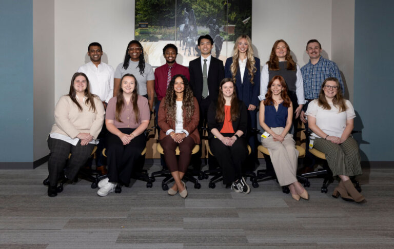 A group of thirteen people who are dressed in business formal attire are standing in two rows, the front row is of six people and the back row is of seven. There is a triptych behind them of a sculpture.