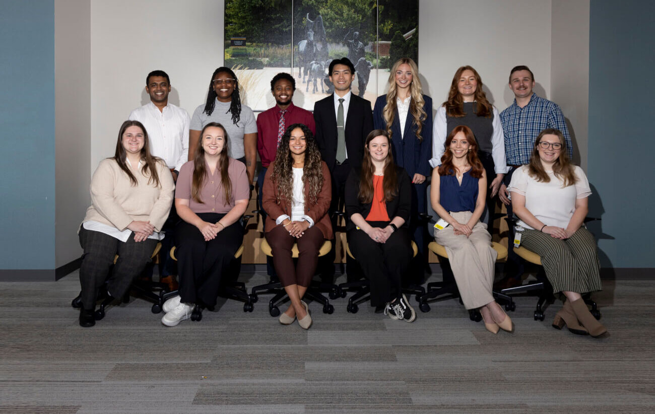 A group of thirteen people who are dressed in business formal attire are standing in two rows, the front row is of six people and the back row is of seven. There is a triptych behind them of a sculpture.