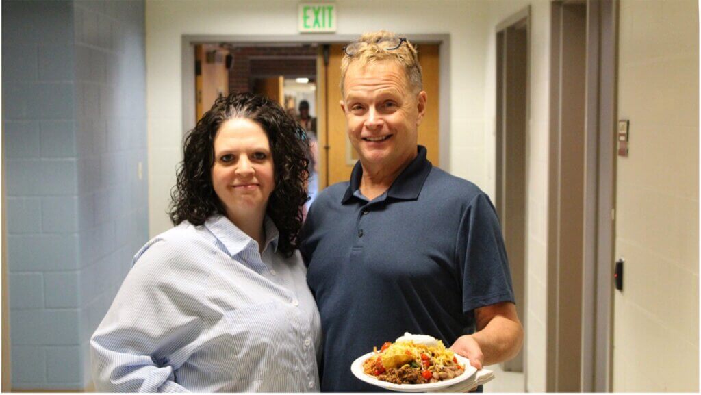 two people are standing next to each other in a corridor, both wearing blue shirts and holding plates of food.