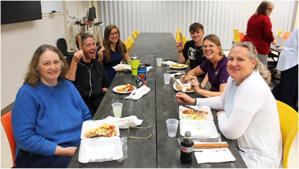 A group of people are sitting at a lunch table, enthusiastically holding up their food items and smiling for a photo. The table has a gray surface and the chairs are yellow and orange.