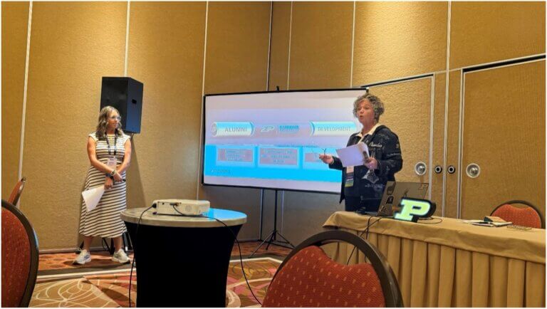 Two people stand on either side of a projection screen. They are giving a presentation. The person on the right appears to be speaking in the photo.