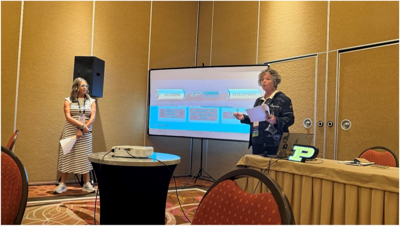 Two people stand on either side of a projection screen. They are giving a presentation. The person on the right appears to be speaking in the photo.
