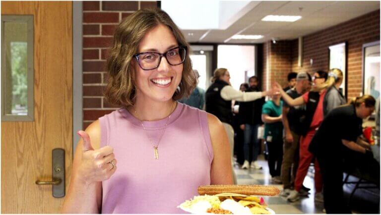 woman in pink shirt is giving a "thumbs up" and is smiling. A group of people are gathered behind her, filling plates with food.