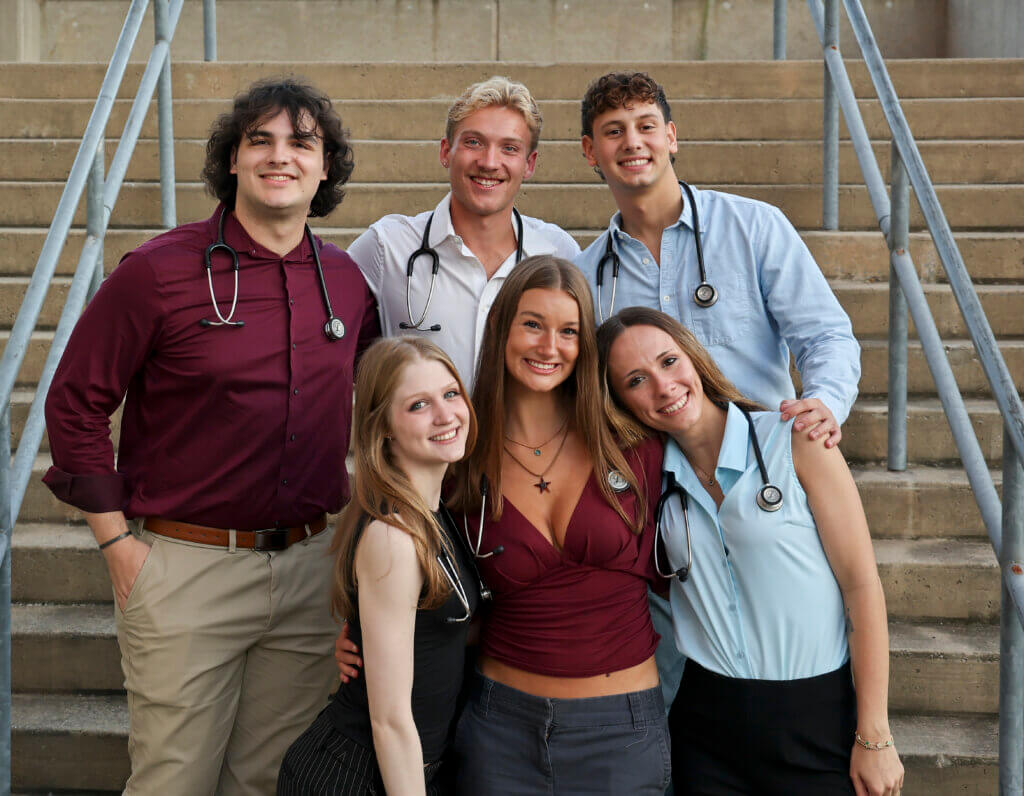 six people are gathered on outdoor steps for a photo. The people are wearing business casual attire and are wearing stethoscopes.