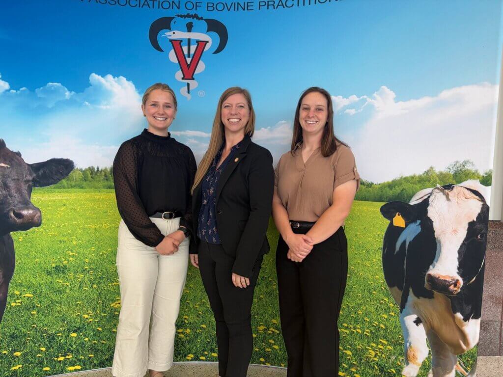 Three people wearing business attire are standing in front of a backdrop image of cows in a pasture.