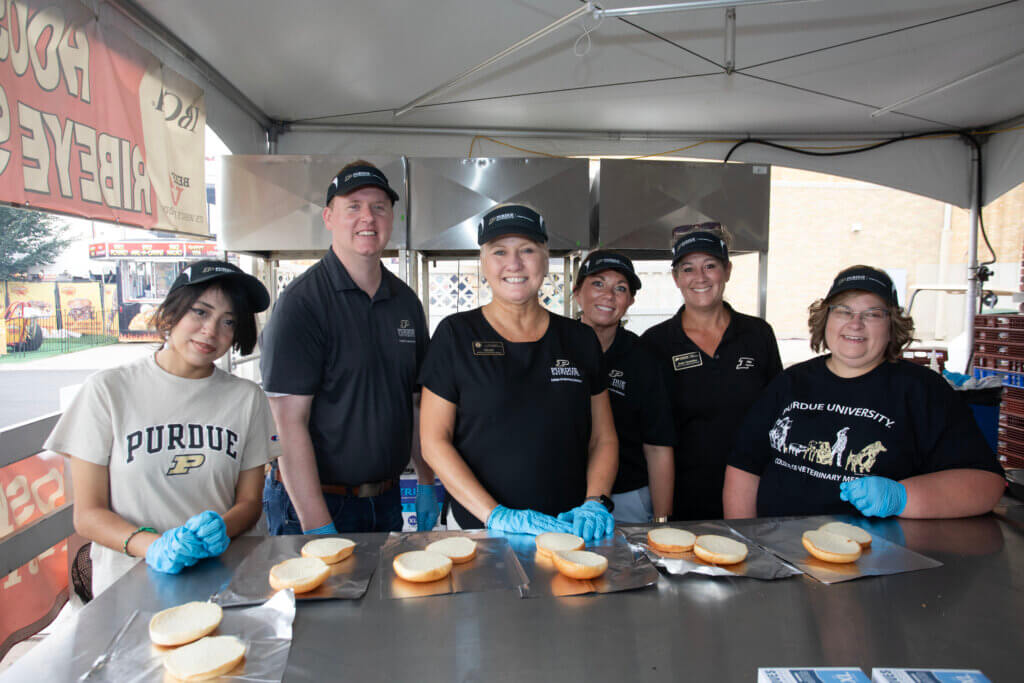 A small group of people appear ready for food preparation. They are standing at a metal table handling bread, and are wearing visors and gloves.