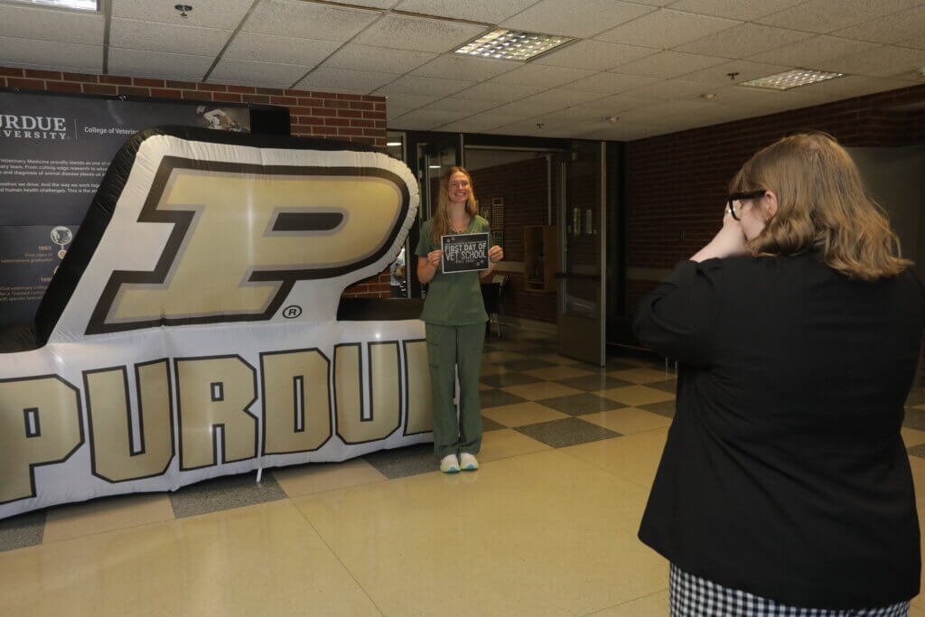A medical student wearing green scrubs is standing in front of a large inflatable Purdue sign, and a person on the right side of the photograph is taking their picture.