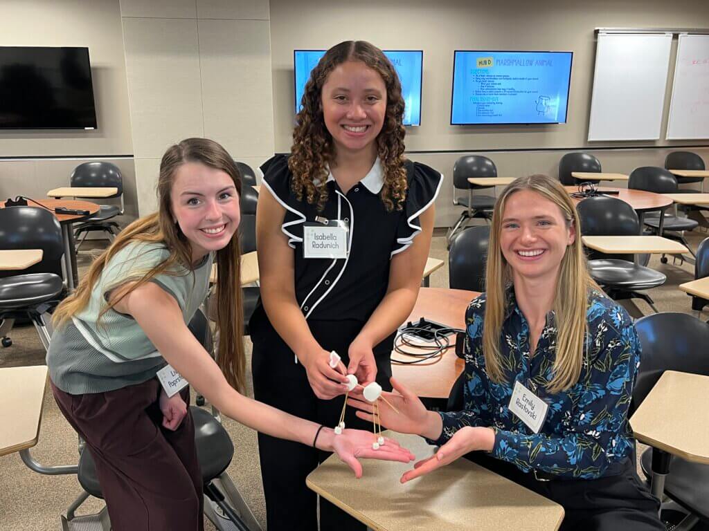 Three people in business casual attire are gathered at a classroom desk and are all holding what appears to be a small sculpture of a dog.