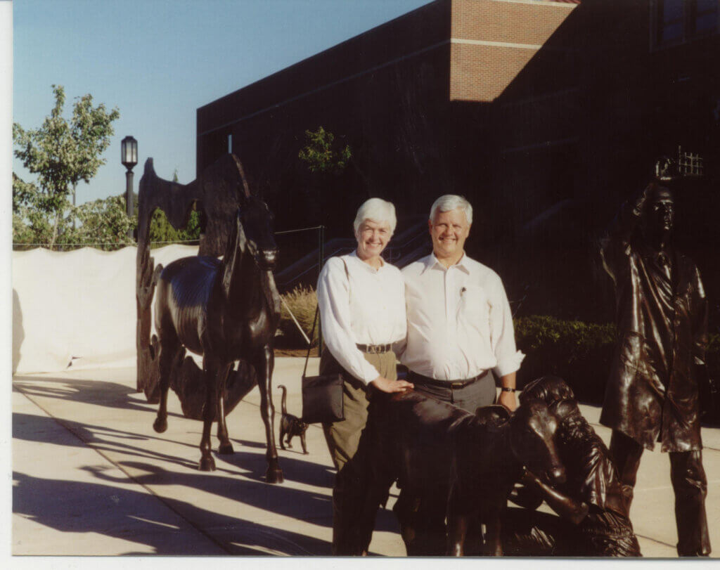 Two people are standing in an outdoor setting, next to a dark-colored statue of an animal. They are both wearing white shirts and are smiling.
