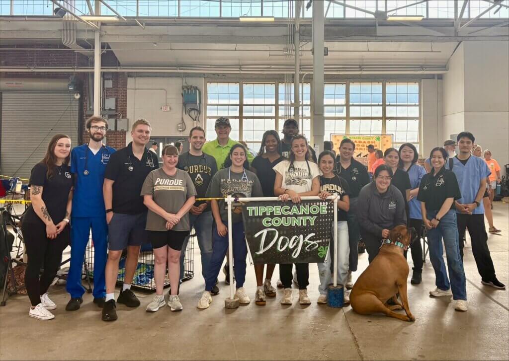A large group of volunteers are posed for a photo, holding a large sign with green and white text, and a dog is in front of the group.