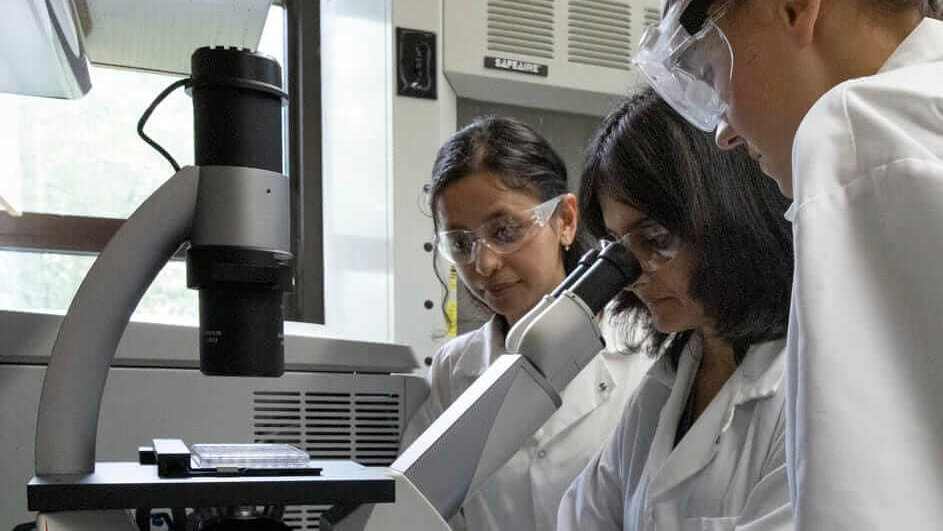 three individuals are standing at a microscope in a laboratory setting. The person in the center is looking into the microscope.