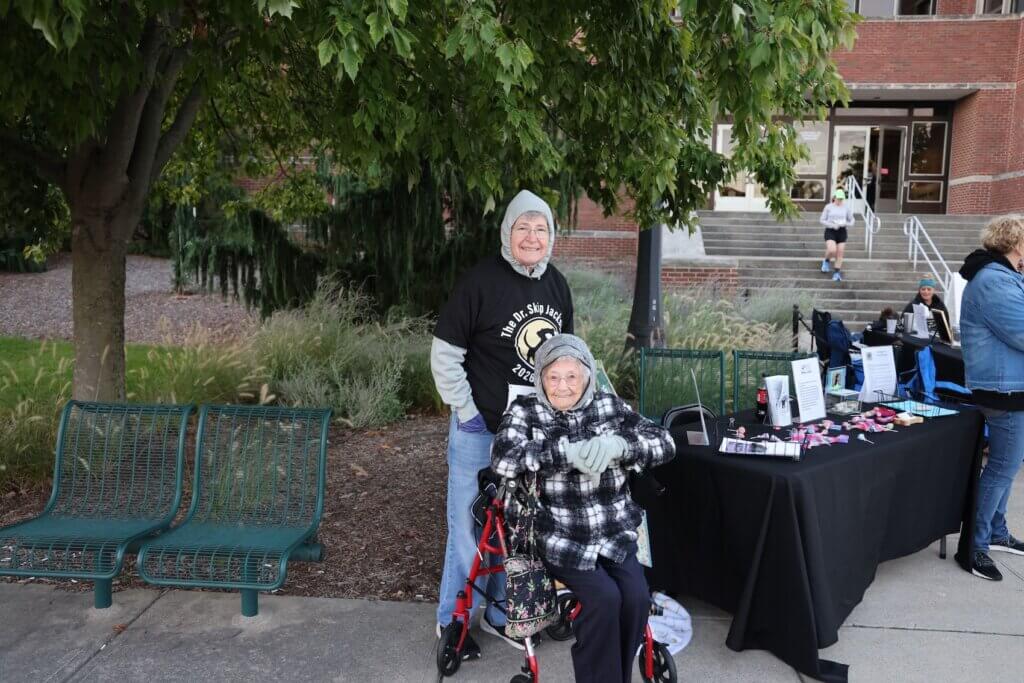 Two people are bundled up in an outdoor setting and are stationed next to a table with merchandise and brochures on it.