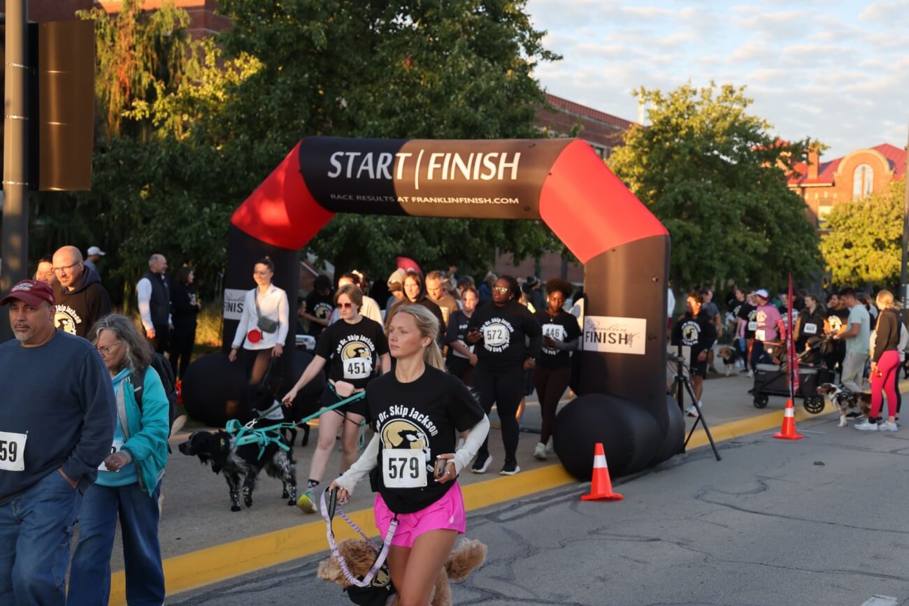 Runners are participating in an outdoor race event on a bright, sunny day. There is a start/finish arch line pictured with several participants under it