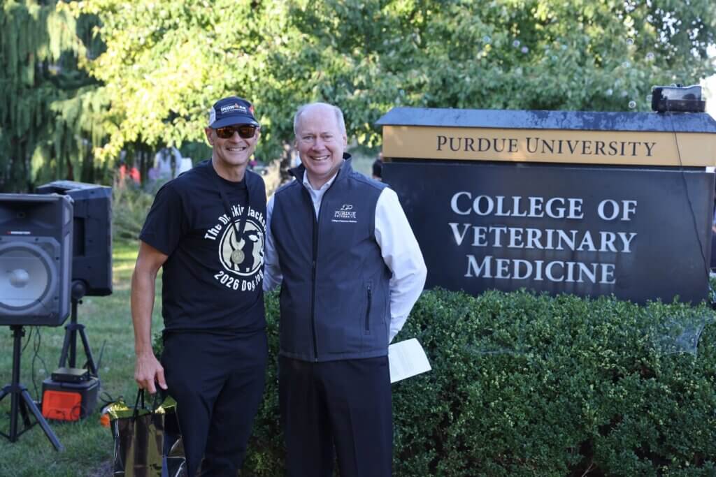 Two people are standing next to a large sign that reads "Purdue University College of Veterinary Medicine". Both people are wearing gray and black clothing, and the figure on the left appears to have just been participating in a sporting event.