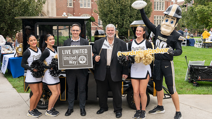 Six figures appear to be posed for a photo celebrating and wearing Purdue University apparel. Purdue Pete, the university mascot, is positioned on the far-right of the group.