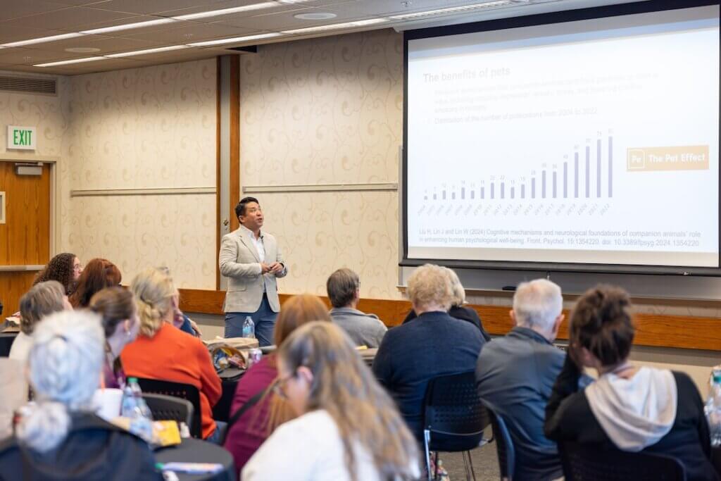 A presentation is taking place in a bright-lit, well-attended room, with the speaker featured in the photo looking at a projector screen.