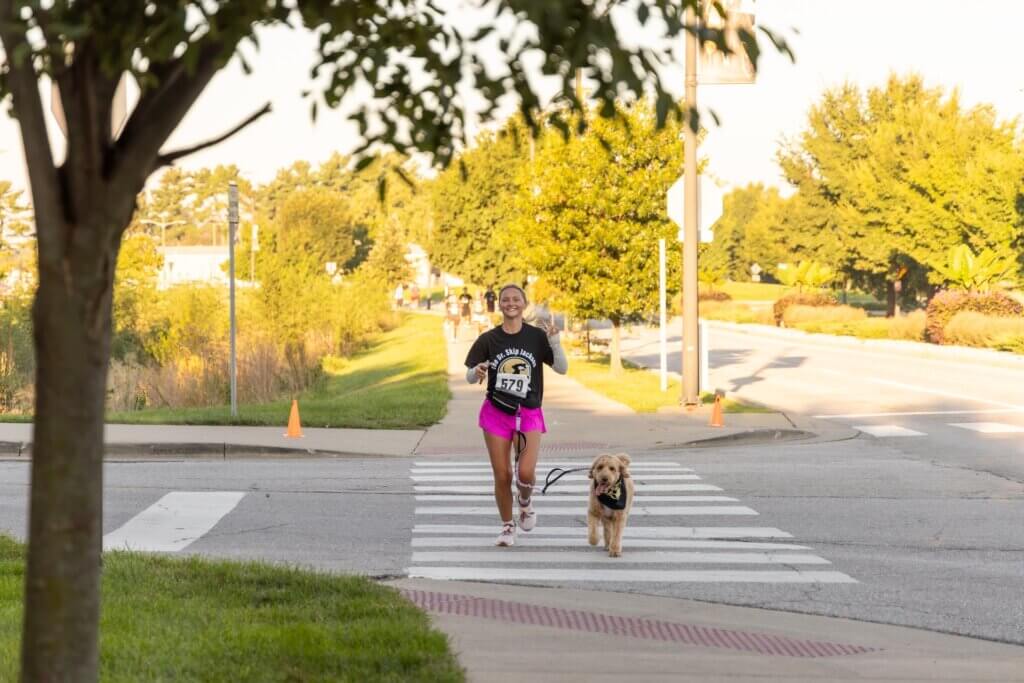 A figure wearing a black shirt and pink shorts is jogging with a dog through an intersection on a bright, sunny day