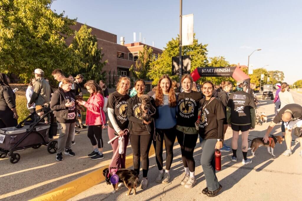 Standing near the start/finish line of a race event, a group of five people are posed with dogs for a photo.