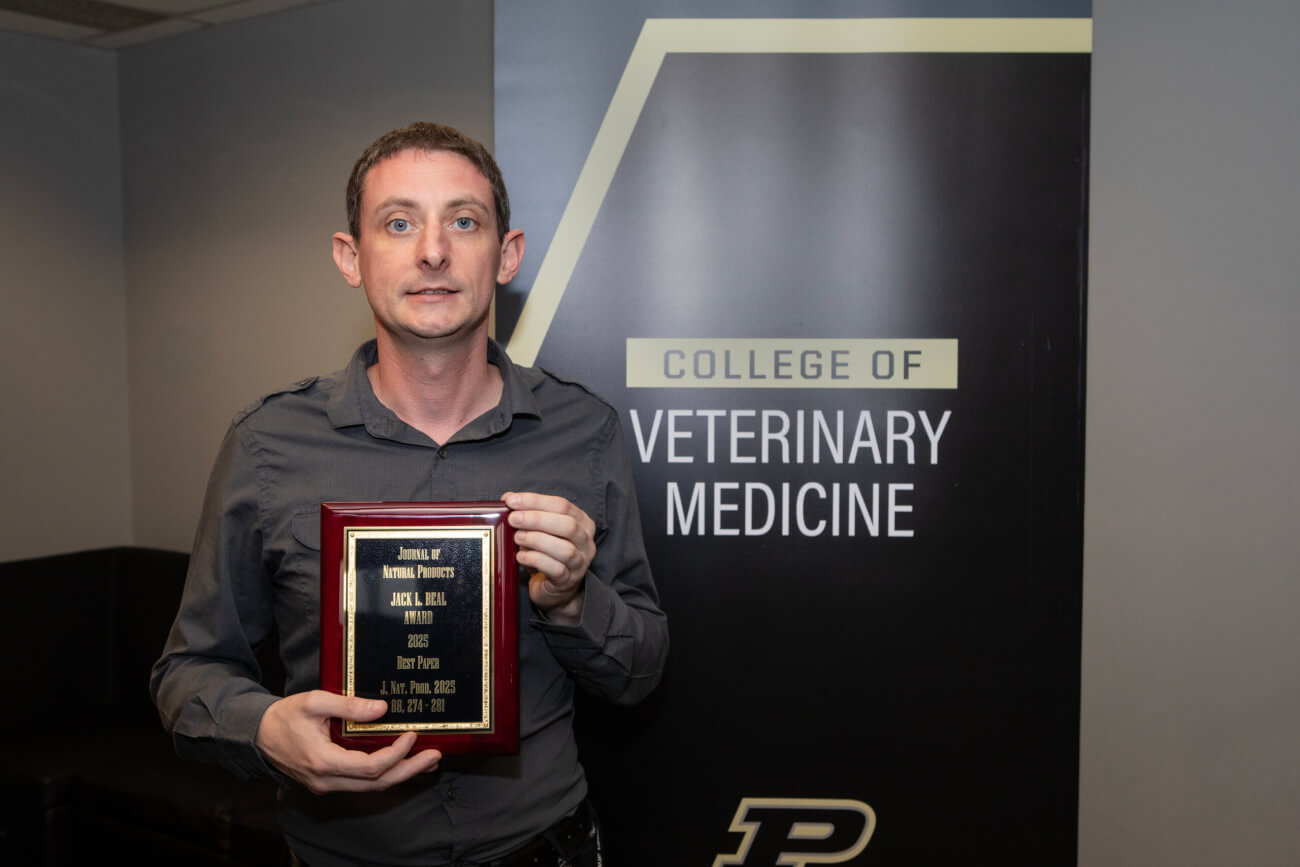 A man is standing in front of a vertical pull-up banner that reads "College of Veterinary Medicine". The man is wearing a gray button-up shirt and holding a plaque award.