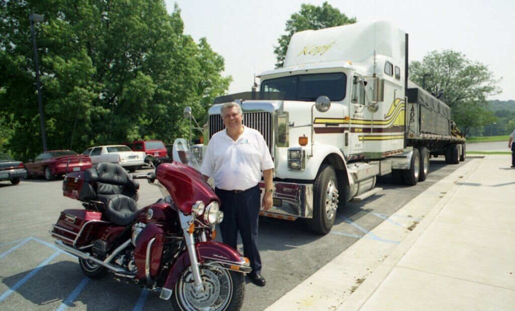 a large semi truck is set in the background with a man standing in front of it. The man is standing next to a maroon-colored motorcycle