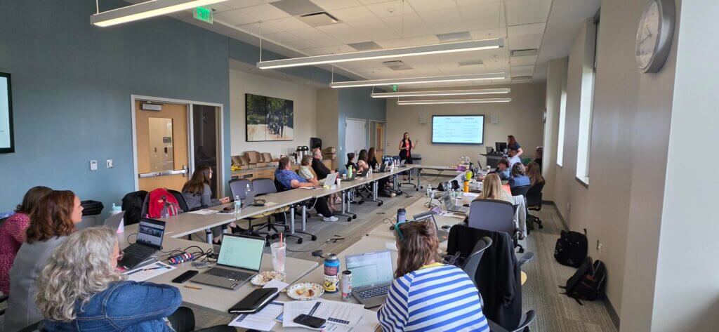 People are sitting around a table with a presentation taking place at the front of the room.