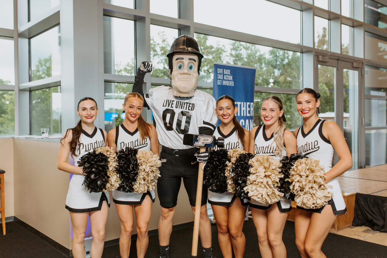 Cheerleaders are gathered with the Purdue University Mascot in front of a window
