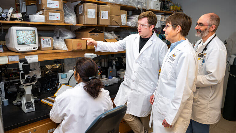 A small group of scientists wearing white laboratory coats are observing a monitor. One of the scientists is pointing at the screen.